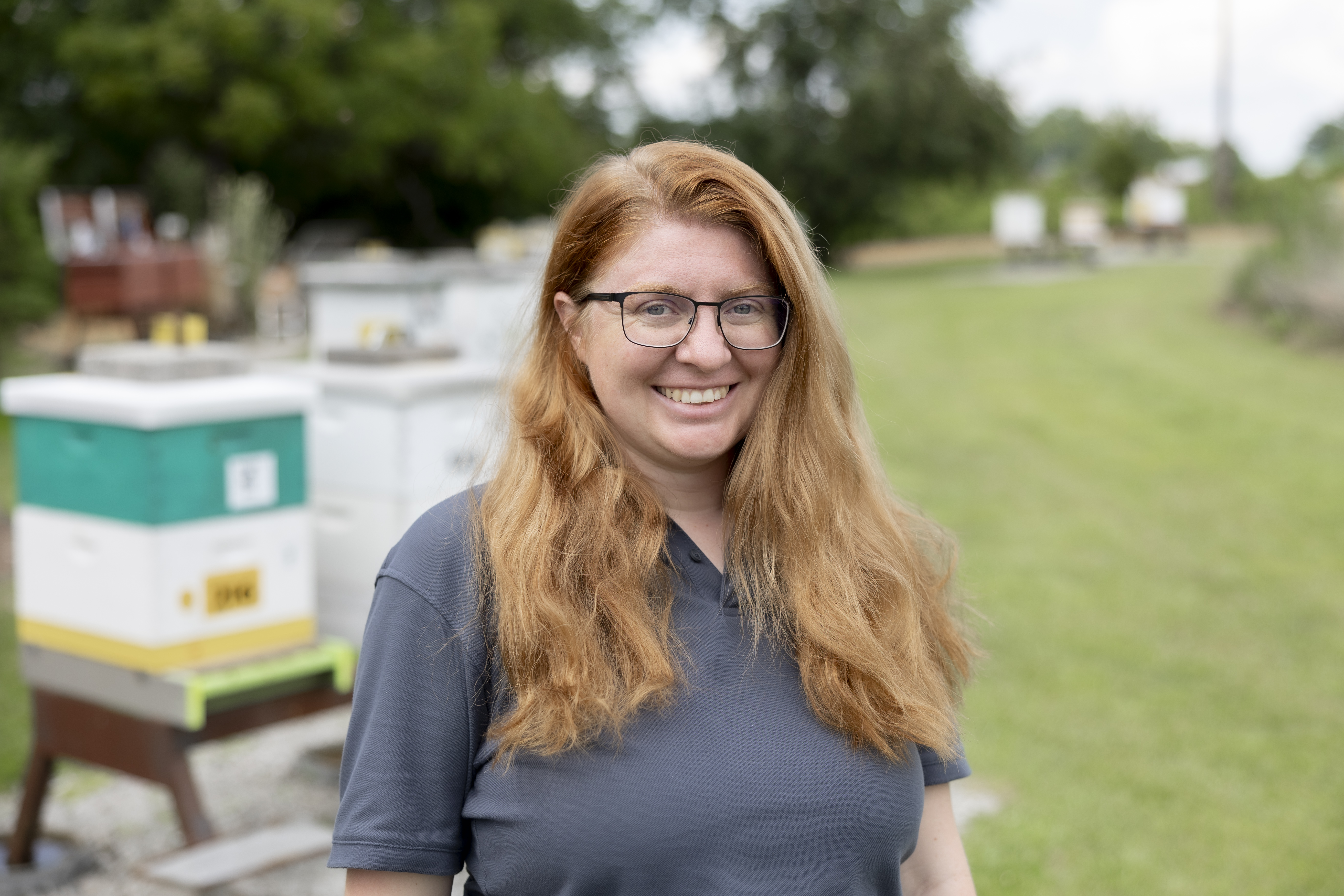 A woman with red hair poses for a headshot with beehives behind her.