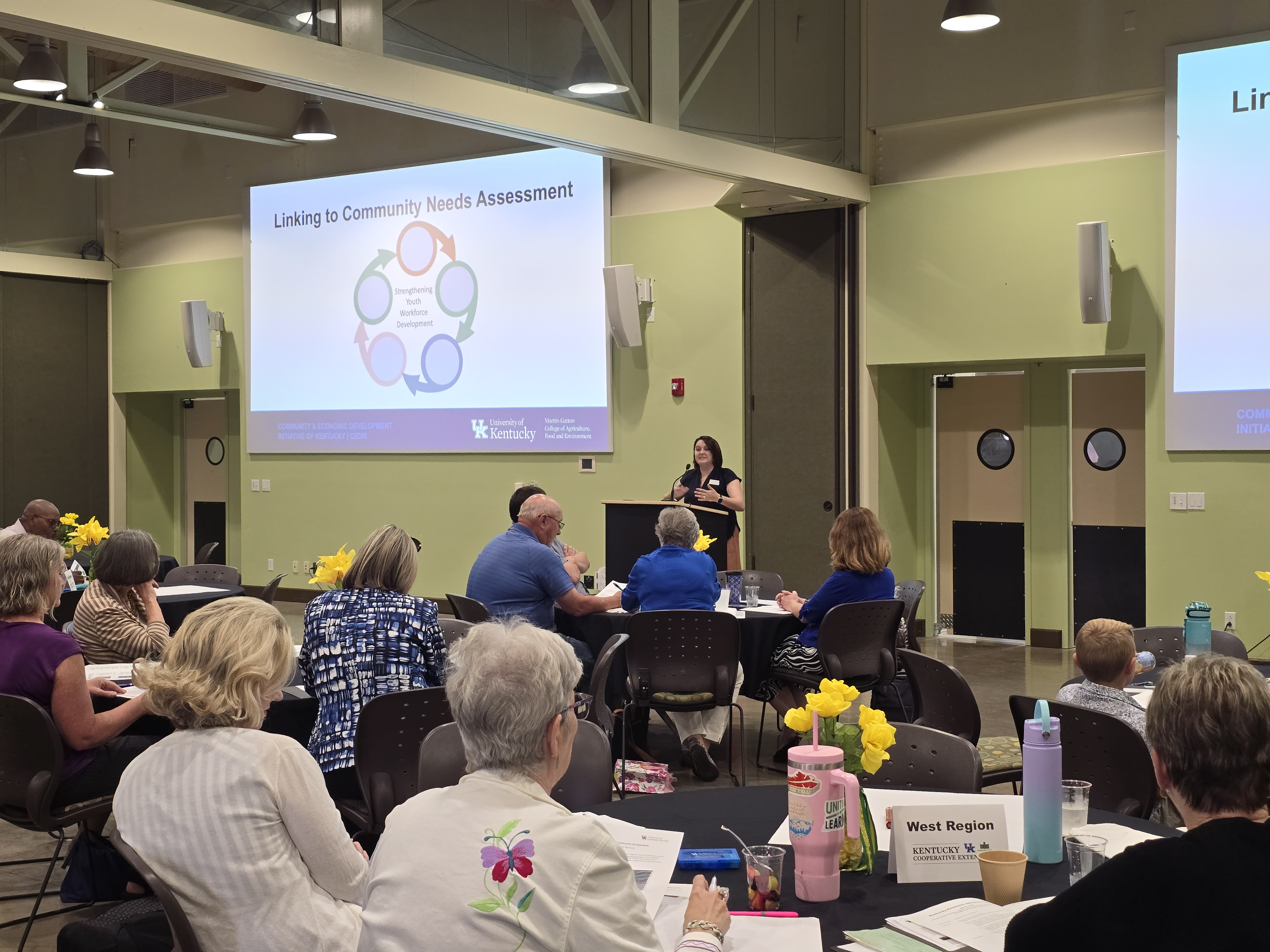 A woman stands at a podium and gives a presentation to a room full of people sitting at tables. The projector screen behind her says "Linking to Community Needs Assessment." 