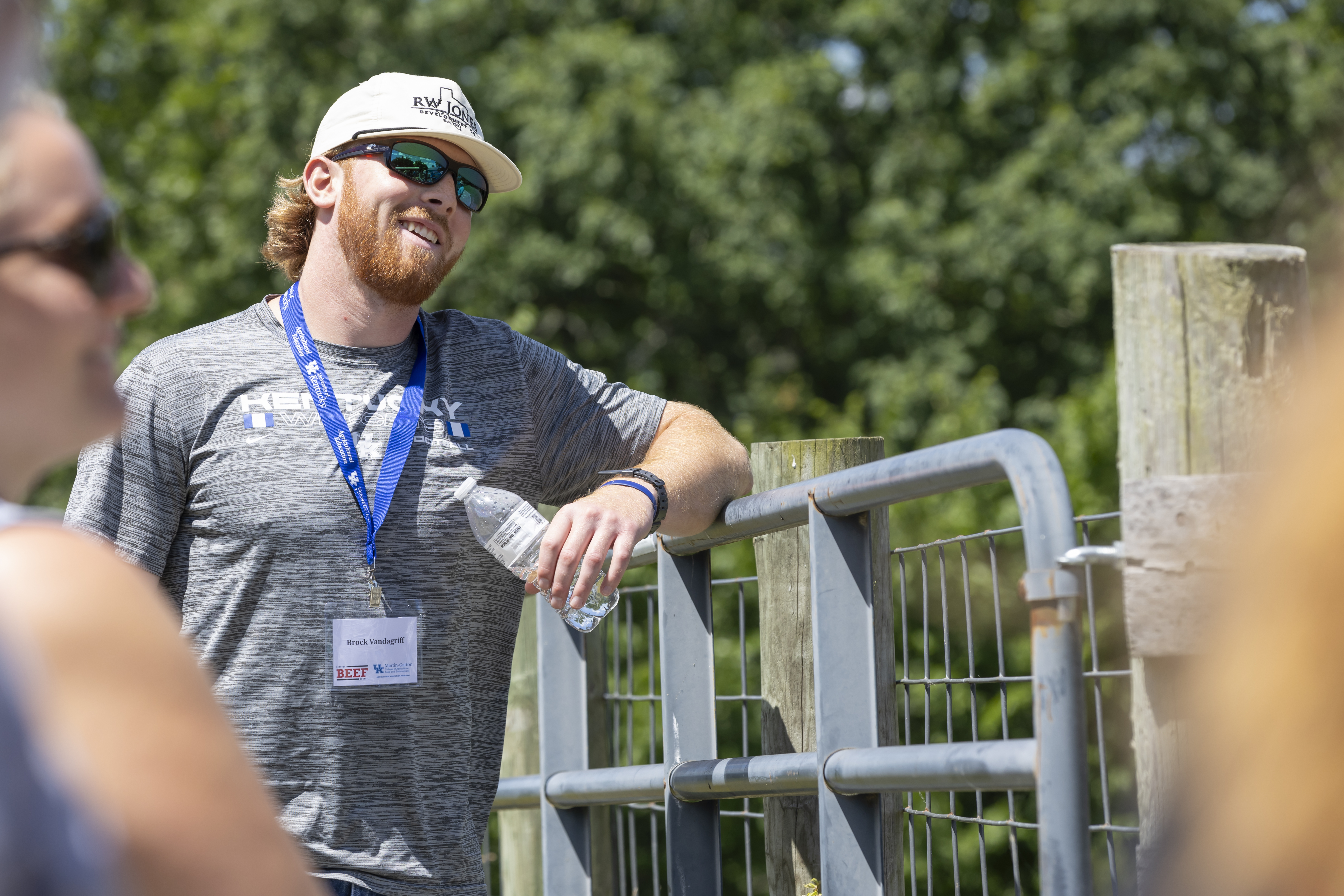 A man wearing a Kentucky Wildcats football shirt leans against a fence and smiles.