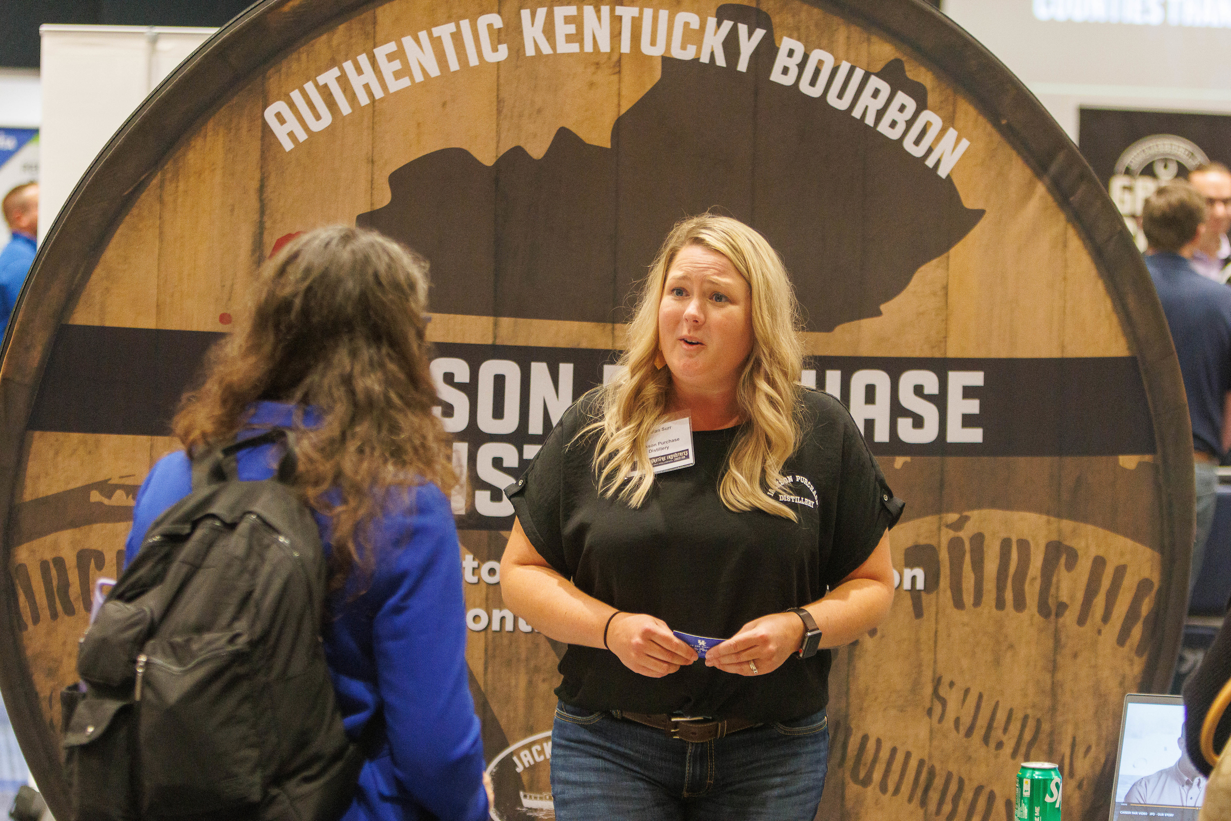 Two women have a conversation in front of a bourbon barrel sign that says Authentic Kentucky Bourbon.