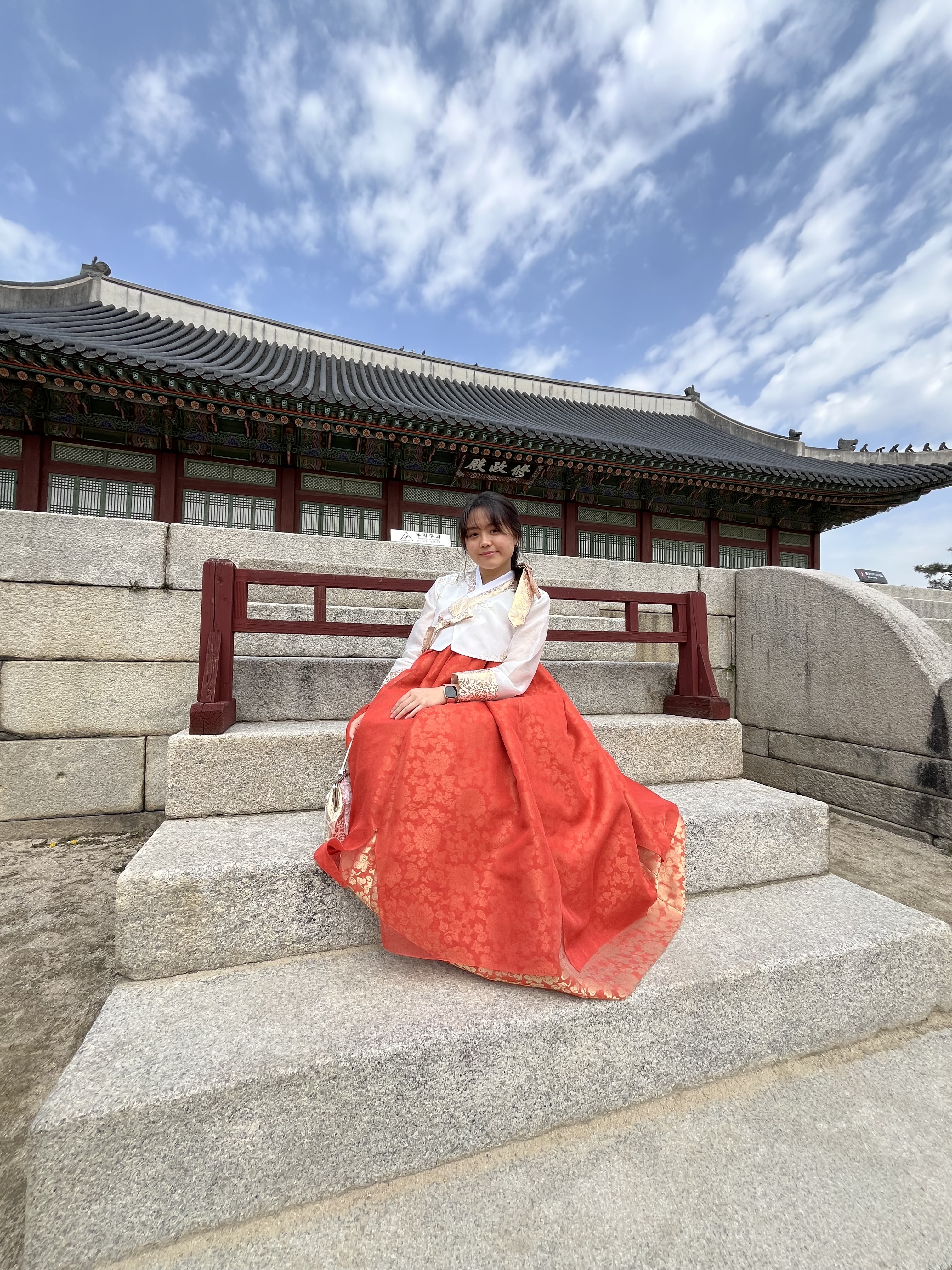 A young Asian American woman wearing a hanbok sits on steps in front of Gyeongbokgung Palace in Seoul, South Korea.