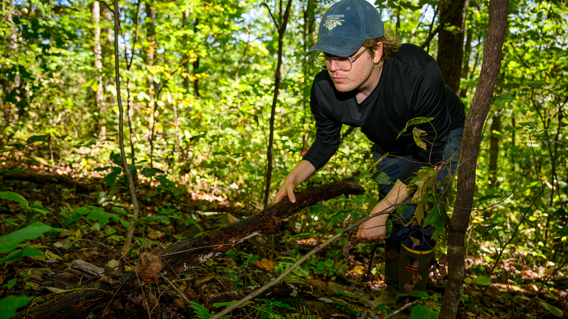 A man wearing a baseball cap lifts up a log to search the underbrush.