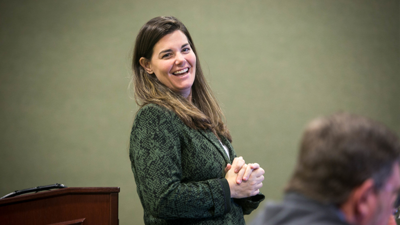 A woman with brown hair and wearing a green jacket smiles as she gives a presentation.