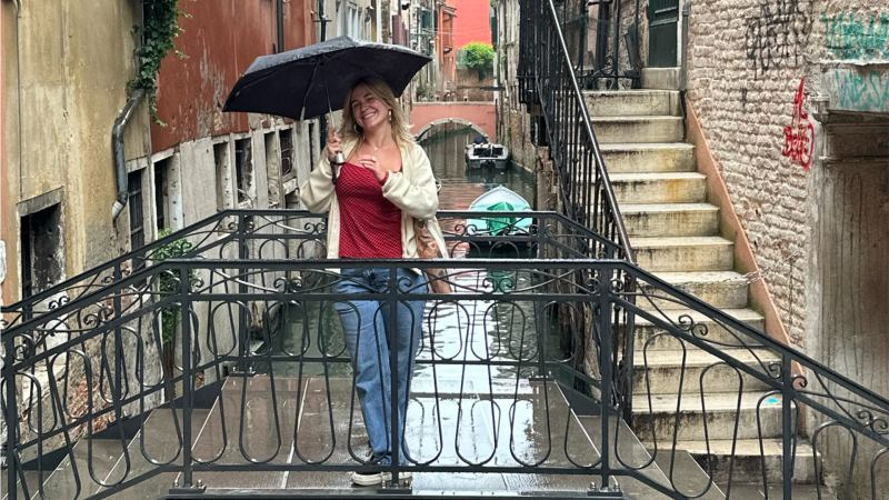 A young woman wearing jeans, a red top and a cream cardigan holds an umbrella while standing on a bridge in Italy.